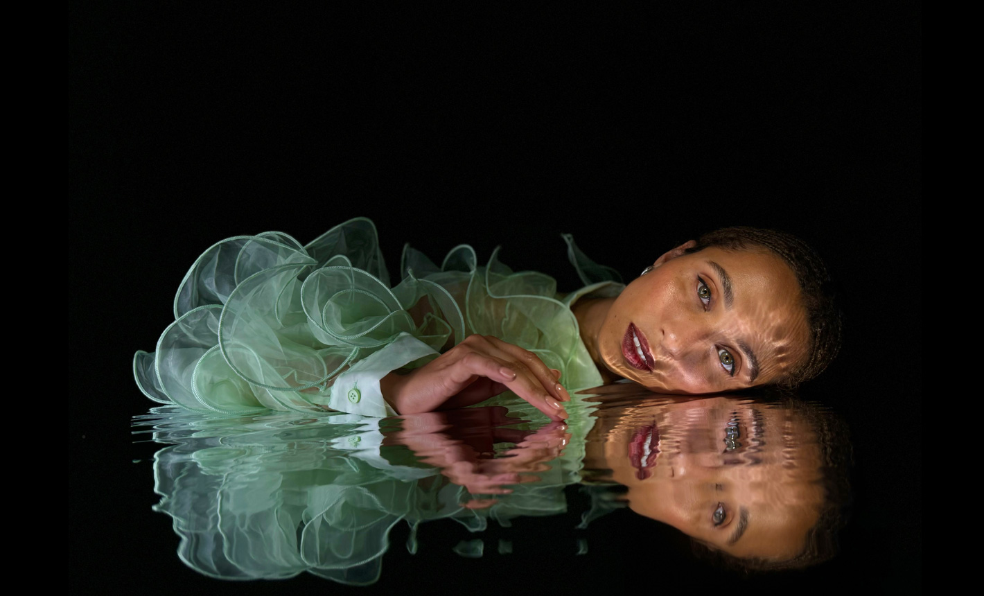 La foto dettagliata, scattata con iPhone 15 Pro, di una donna china su uno specchio d’acqua increspata su cui si riflette il suo viso, su uno sfondo nero
